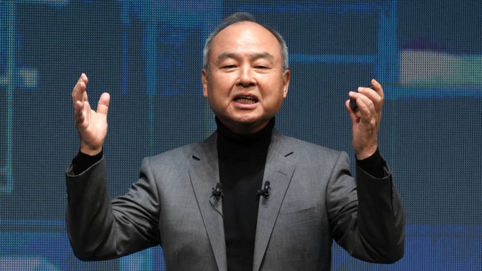 Masayoshi Son, wearing a gray suit and black turtleneck, speaks with his hands raised