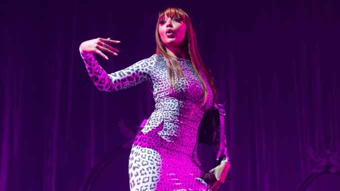 A female singer in a tight-fitting leopard-print dress performs on stage, wearing a handbag over one shoulder and extending her other arm