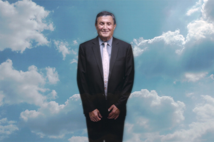 A man in a dark suit stands in front of background showing a blue sky and clouds