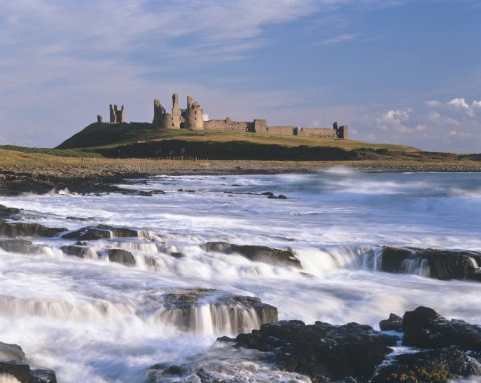 The ruins of the 14th-century Dunstanburgh Castle in the distance, with the sea and a rocky beach in the foreground