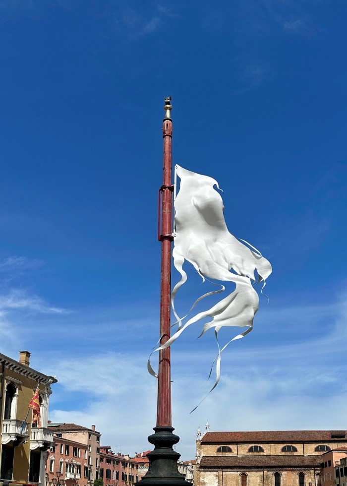 A flag-like white sculpture hangs half-mast on a tall, red pole against a vividly blue sky and pastel-shaded buildings