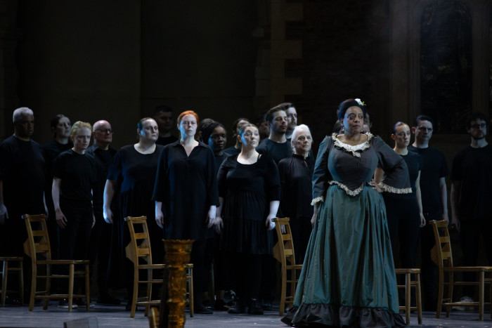 A serious-looking woman wears a drab dark grey 19th-century dress as she stands in front of a chorus clad in black