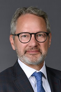 A man with glasses and greying hair, wearing a suit and tie, looks into the camera