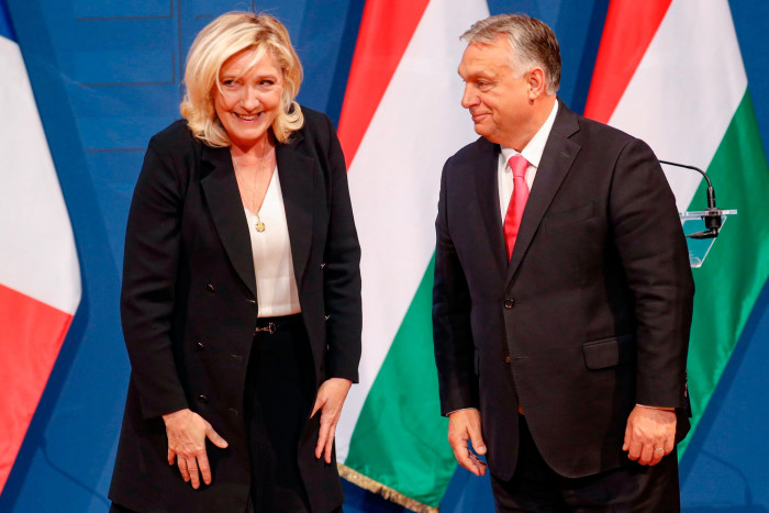 A woman in a dark suit stands beside a man in a dark suit in front of three Italian flags