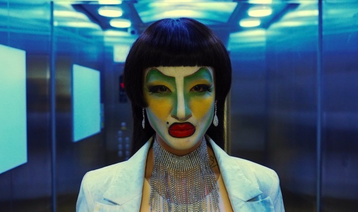 A person in a mirrored lift with a black bob wig and white face makeup with green eyes and yellow cheeks