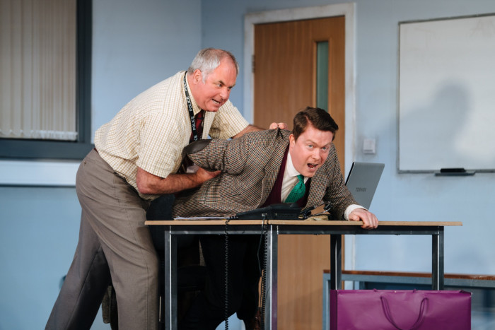 In an office, a man stands and pushes another man over a desk while twisting his arm behind his back