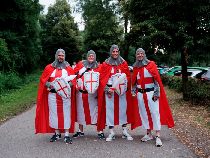 Four smiling men stand on a tree-lined path. They are wearing medieval knight’s outfits, with fake chainmail helmets, red cloaks and white robes marked with a large red cross