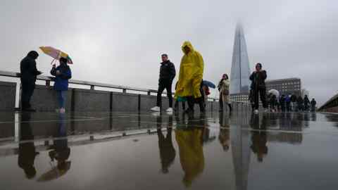 Commuters in raincoats and carrying umbrellas are reflected in rain on London Bridge with a view of the Shard in the background