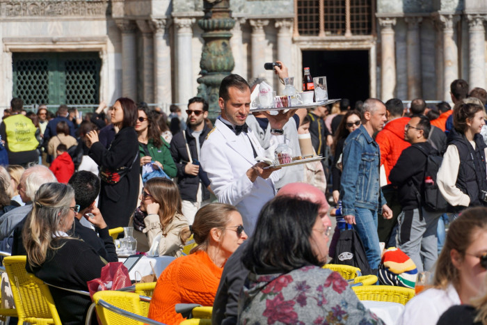 A waiter in a white jacket balances two trays of drinks next to tables where people are seated. Behind him walk crowds of people