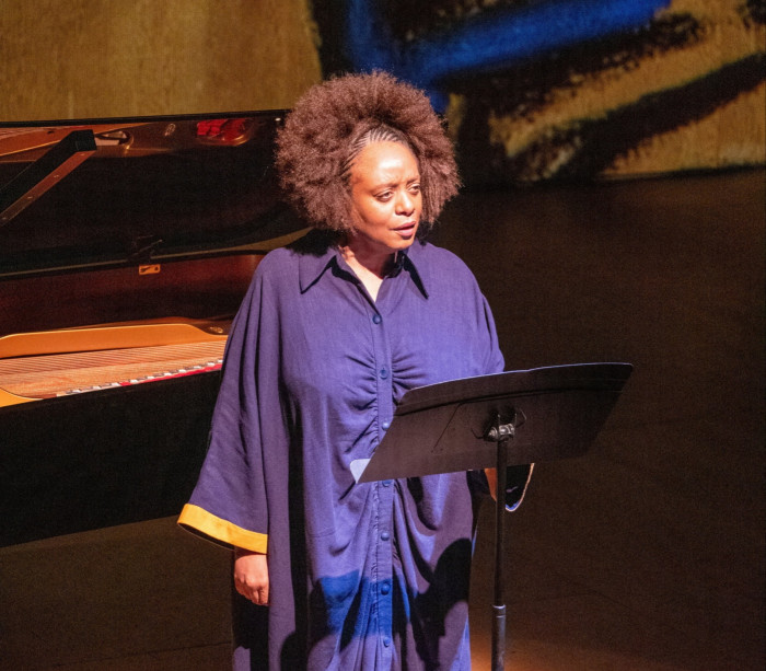 A woman stands singing on stage with a music stand in front of her; behind her are projections of abstract artworks