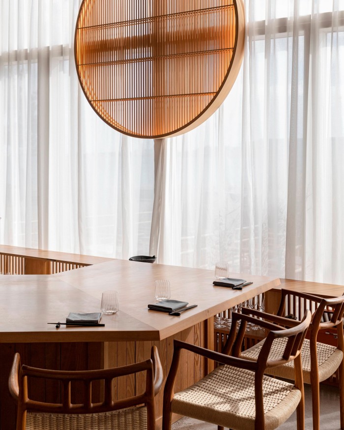 Wooden chairs at the counter of Sushi Anaba, behind which a large slatted wooden circle hangs from the ceiling in front of net-curtained windows