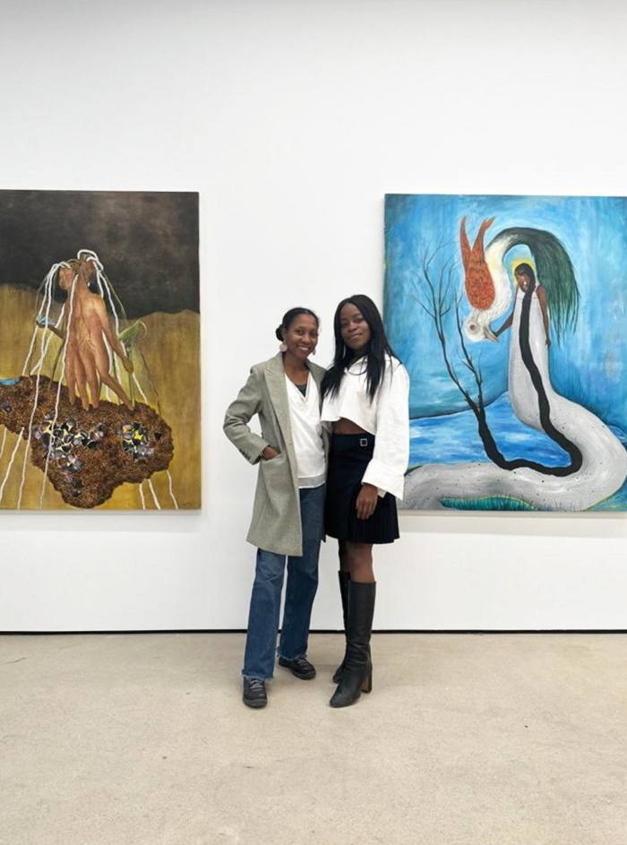 Taiye Idahor and Aindrea Emelife pose in front of paintings on a wall