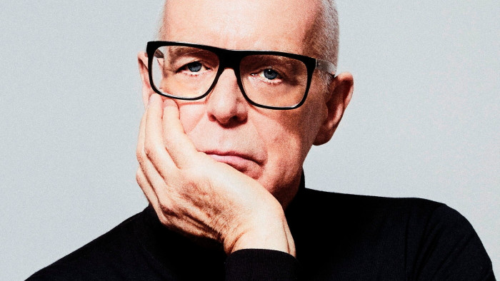 Neil Tennant, with white hair and black frame glasses and wearing a black turtleneck, poses unsmiling with his hand under his chin