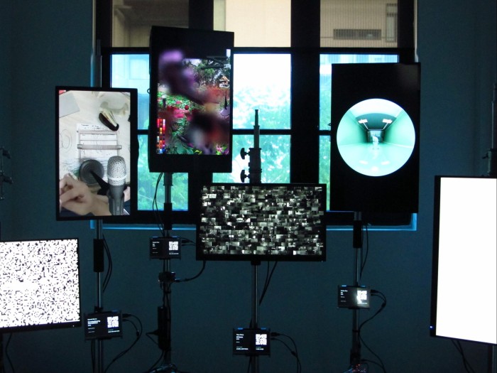 Several digital screens are positioned at various heights in front of the window of a gallery room