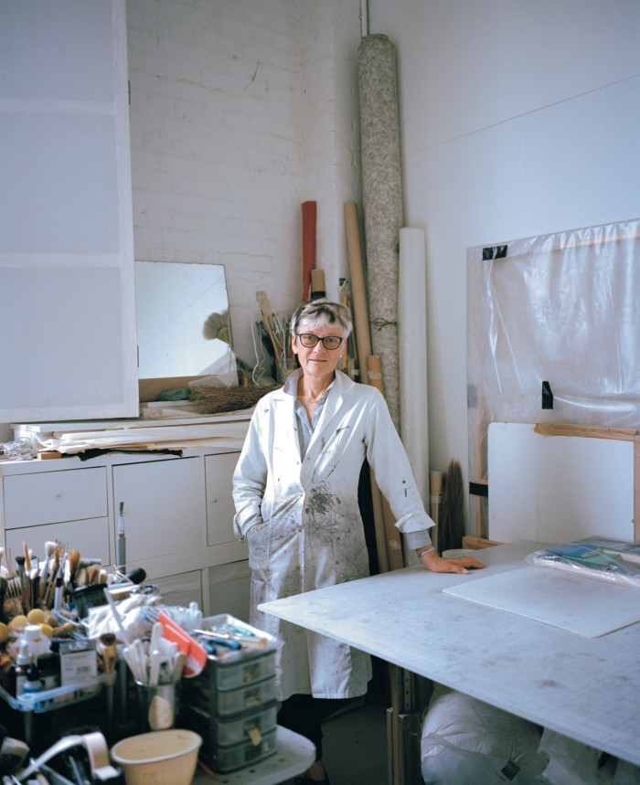 A middle-aged woman dressed in a white shirt and wearing dark glasses stands in a naturally lit studio filled with brushes and canvases while smiling at the camera. 
