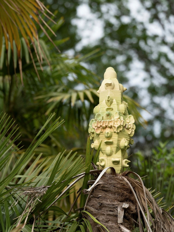 A light green, totem-like small sculpture rises on top of a palm tree against a blurred, vegetation-filled background 