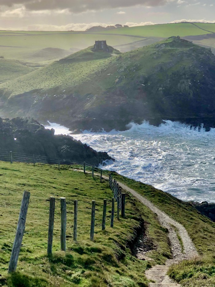 A path leading down a hill to a choppy sea at Port Quin, an inlet on the Cornish coast. On the other side of it are green hills and fields 