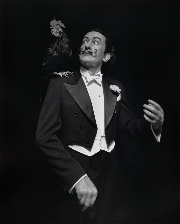 In a monochromatic photograph, a moustached middle-aged man wearing a tailored suit stares surprised at the chicken resting on his right shoulder