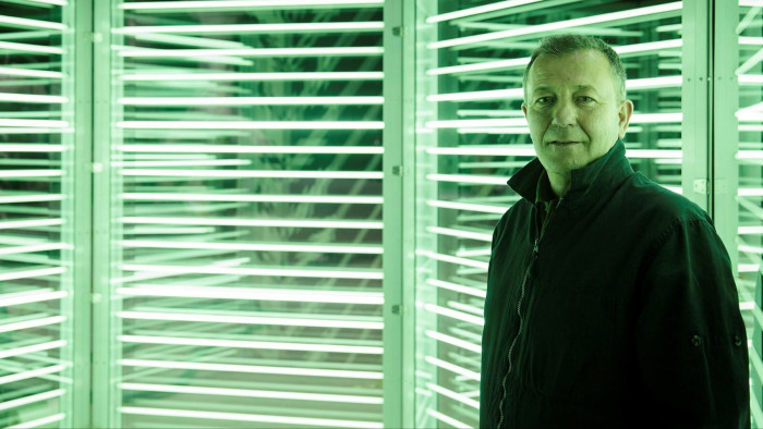A middle-aged man wearing a dark bomber jacket looks at the camera against a green neon-lit background