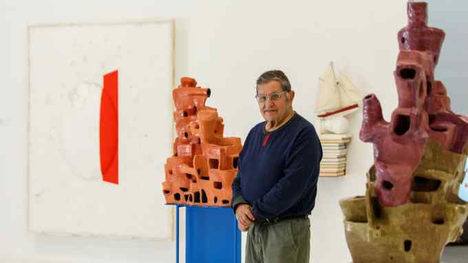 Photograph of a man in glasses standing between two ceramic sculptures, a painting and a small wall sculpture behind him, taken in a gallery