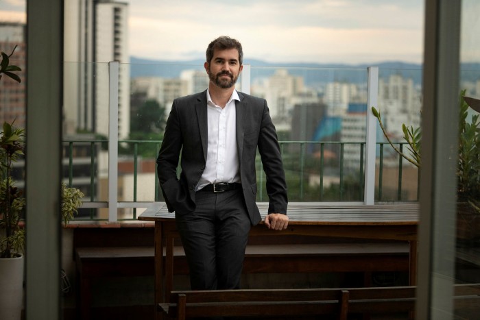 Luiz Amaral wearing a suit against a city landscape by a window