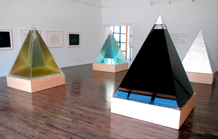 Four pyramidal, reflective sculptures in green, black, light blue and silver, respectively, are shown in a gallery alongside wall art