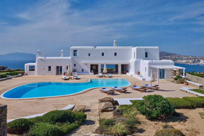 a large white traditional L-shaped villa with small windows built around a large pool and terrace