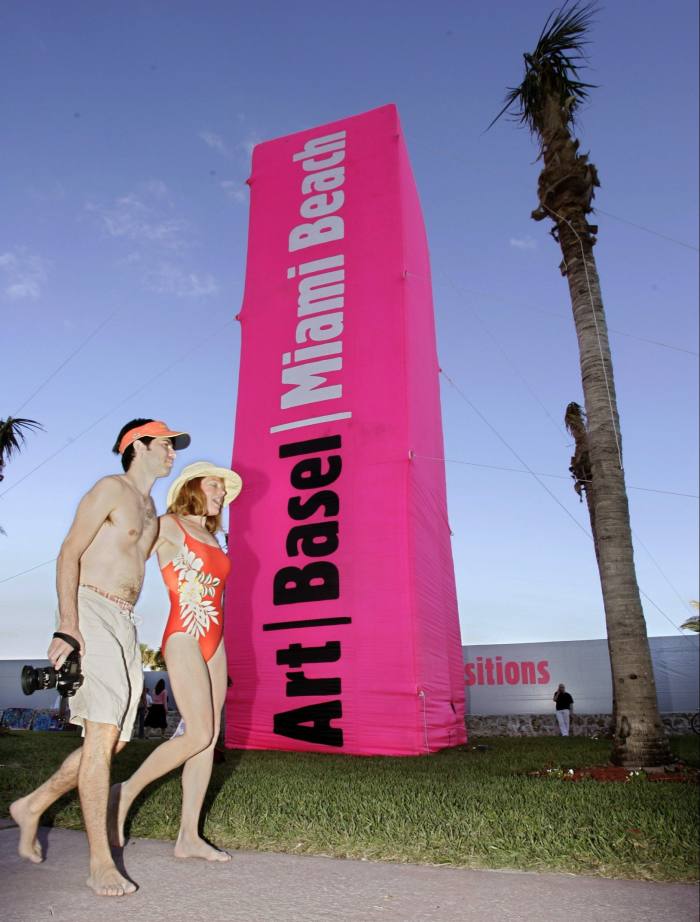A man and woman wearing swim suits walk past a pink Art Basel advertising column protruding from the grass verge 