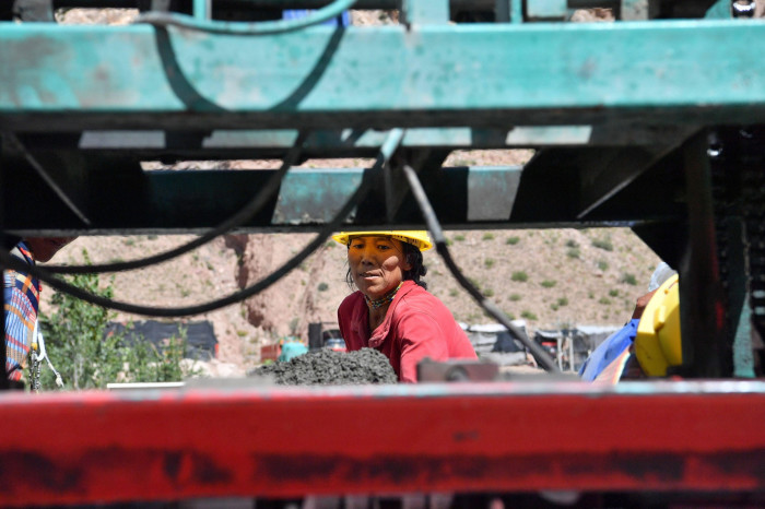 A construction worker, donning a yellow hard hat and red shirt, is framed by industrial machinery as she works at a construction site. The background features a rocky landscape and other workers in safety gear