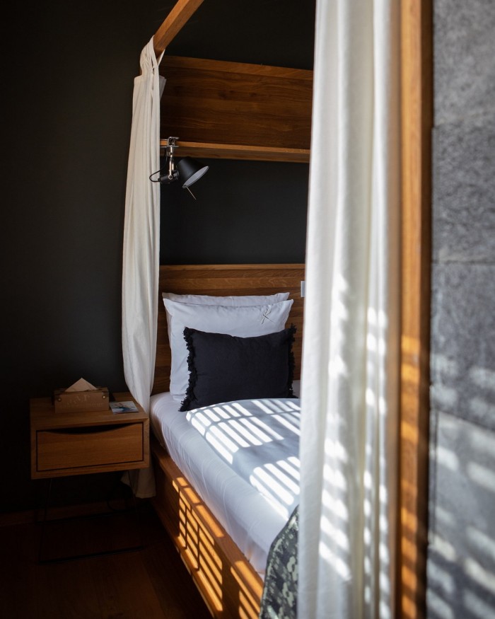 A pale-wood bed onto which the sun is shining through venetian blinds in Bryggen Guldsmeden’s 211 rooms