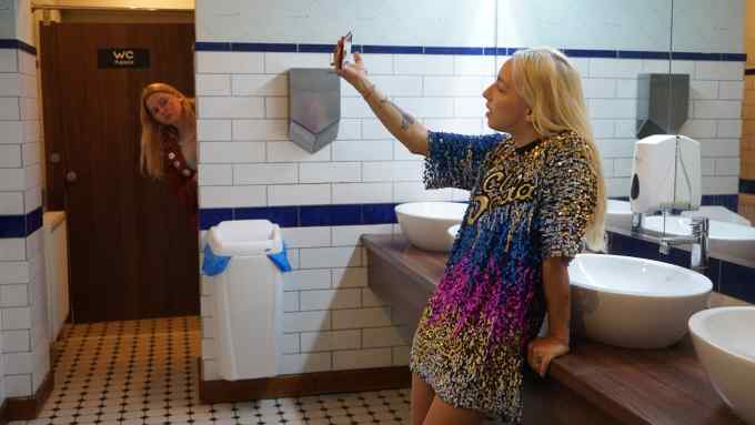 Young blonde-haired woman in public bathroom holding mobile phone up to take a selfie as another woman peeks at her around a wall in the background