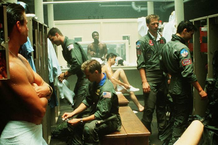 Pilots in a locker room, some in uniform, some of them bare-chested and with towels around them
