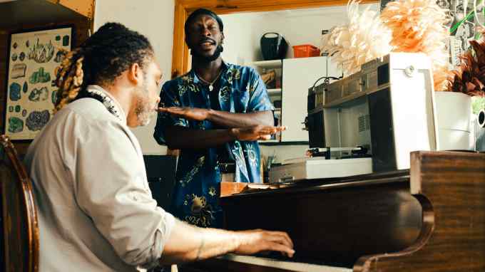 Producer Neue Grafik wears a grey shirt and plays the piano next to Brother Portrait who raps/sings in a blue shirt and yellow patterns in a room in which a record player, feather dusters and a record player are visible