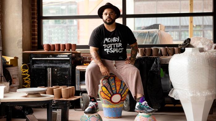 A young man in a black hat and black T-shirt which reads “Money, Potter, Respect” sits on a brightly coloured vase