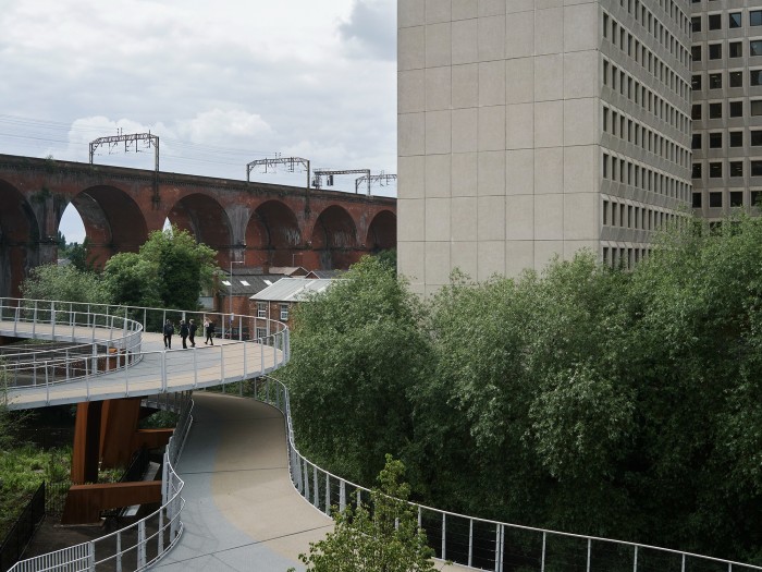 Four people walk up a modern curved overpass. A historic viaduct is in the background