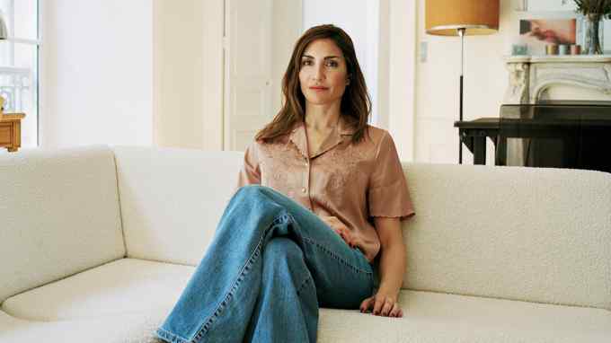 A woman with long brown hair wearing a silk shirt and blue jeans sits on a cream sofa with her legs crossed