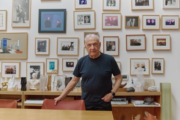 Gehry stands in his Los Angeles studio in front of a wall of photographs
