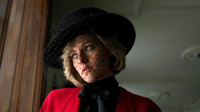 A woman with short blonde hair wearing a red coat and black hat with a net veil looks forlornly out a window