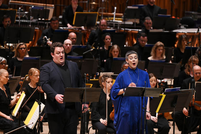 A male and a female singer stand performing in front of an orchestra