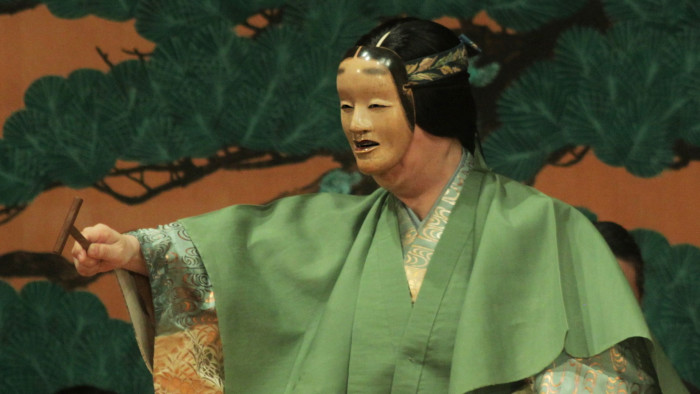 An actor wearing a Japanese noh theatre mask and a green gown performs in front of a backdrop of stylised trees against a red sky