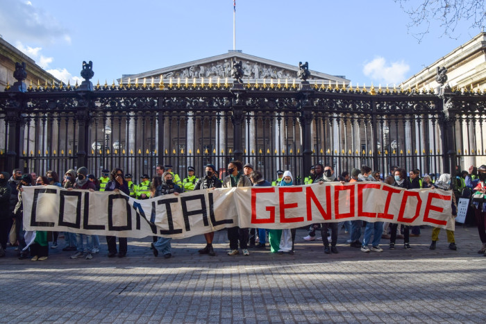 People outside the gates of a very large columned building hold up a sign that says ‘colonial genocide’