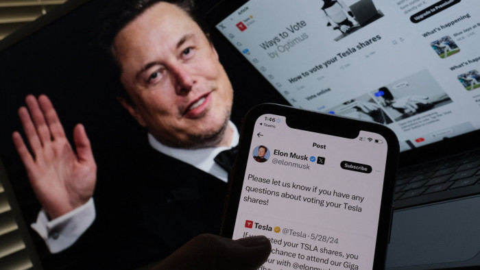 An image showing Elon Musk’s campaign on the social media platform X on a smartphone, and an image of Musk waving on a screen behind