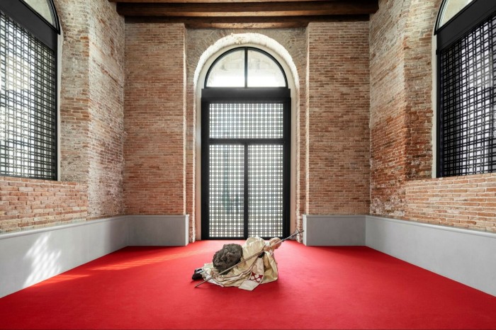 A sculpture resembling Pope John Paul II lies on a red carpet in front of a large steel gallery door