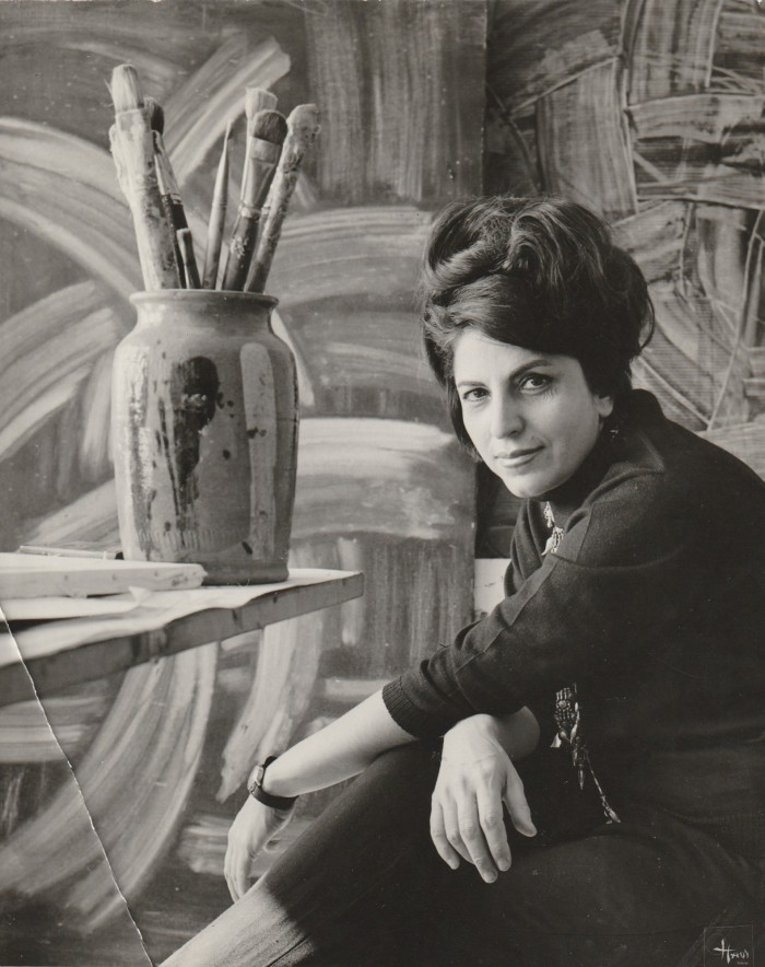 Black and white photo of a woman with big hair in her studio in front of a big painting