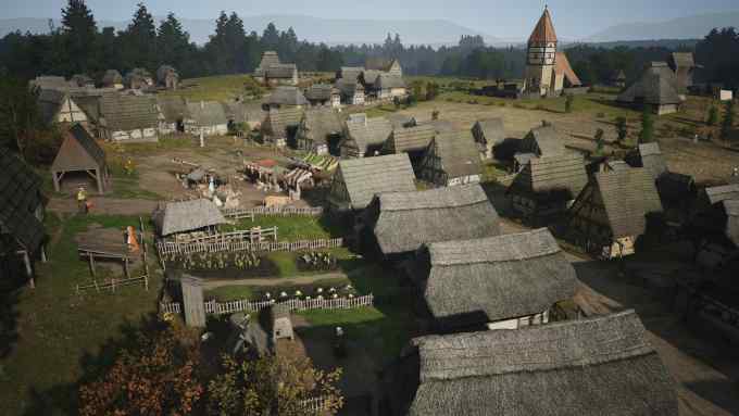 In an image from a video game, a medieval village is seen from above, with thatched roofs, smallholdings and a church, with hills in the distance