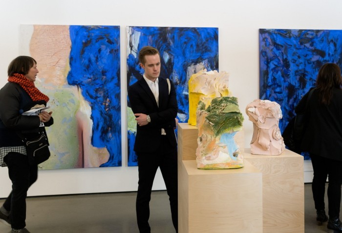 A man in a black suit looks at some large abstract works which appear to be sculpted in clay then brightly painted