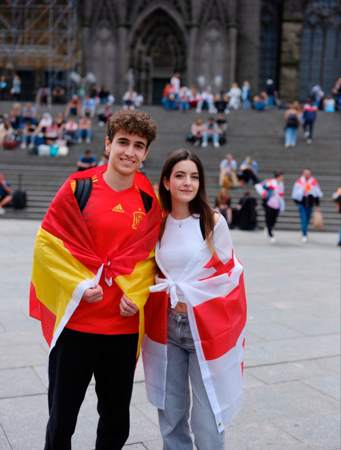 A young couple, smiling, stand in front of the steps of a cathedral. He wears a red football shirt and has the yellow and red Spanish flag draped over his shoulders. She wears a white t-shirt and has the red and white flag of George around her