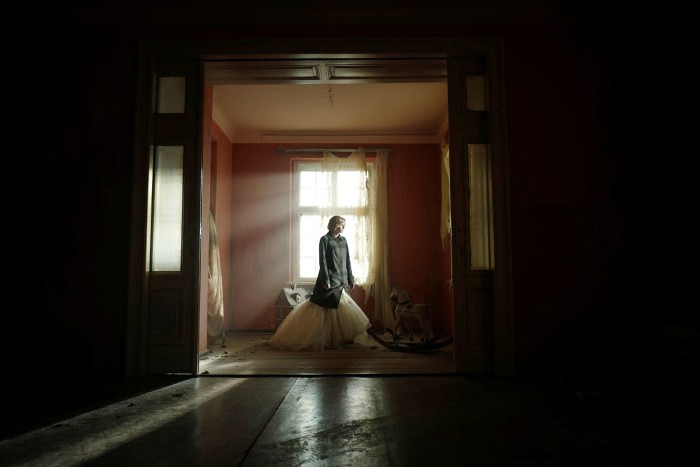 A woman in a coat and ballgown wanders through an old nursery of a dimly lit, dilapidated house