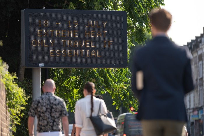 Passersby walk towards an LED sign saying “18 -19 July extreme heat only travel if essential”