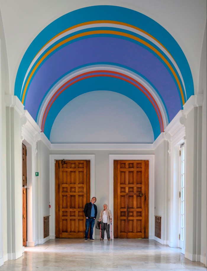 A man and a woman stand looking up at a striped ceiling mural 